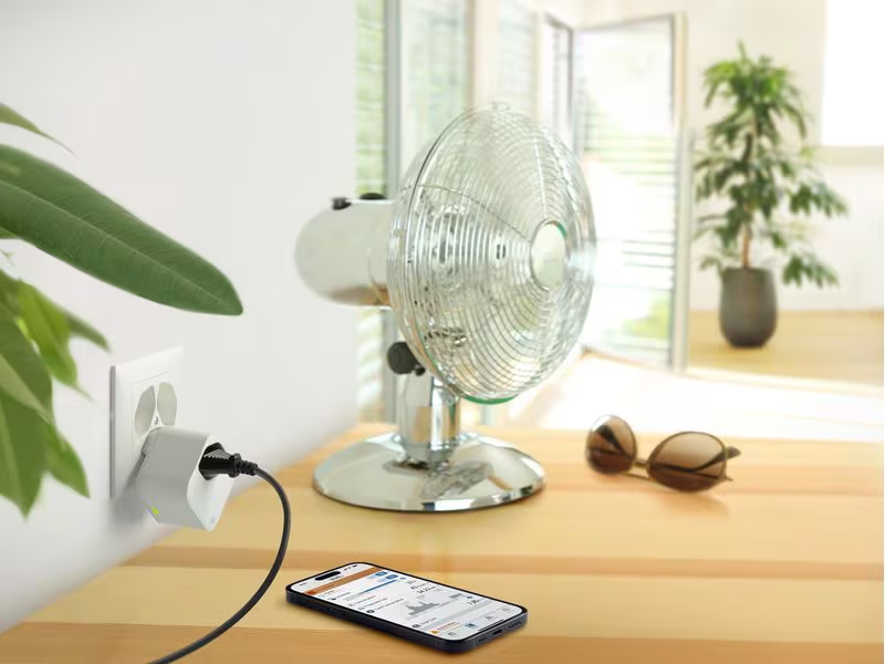 Ein silberner Ventilator steht auf einem Tisch. Ein Handy und eine Sonnenbrille liegen daneben.