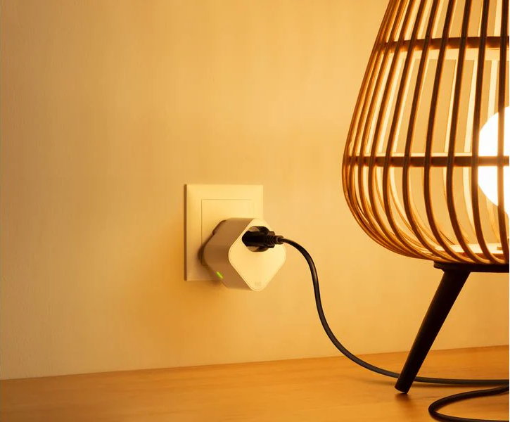 Ein weißer Smart Plug steckt in einer Steckdose neben einer Lampe.