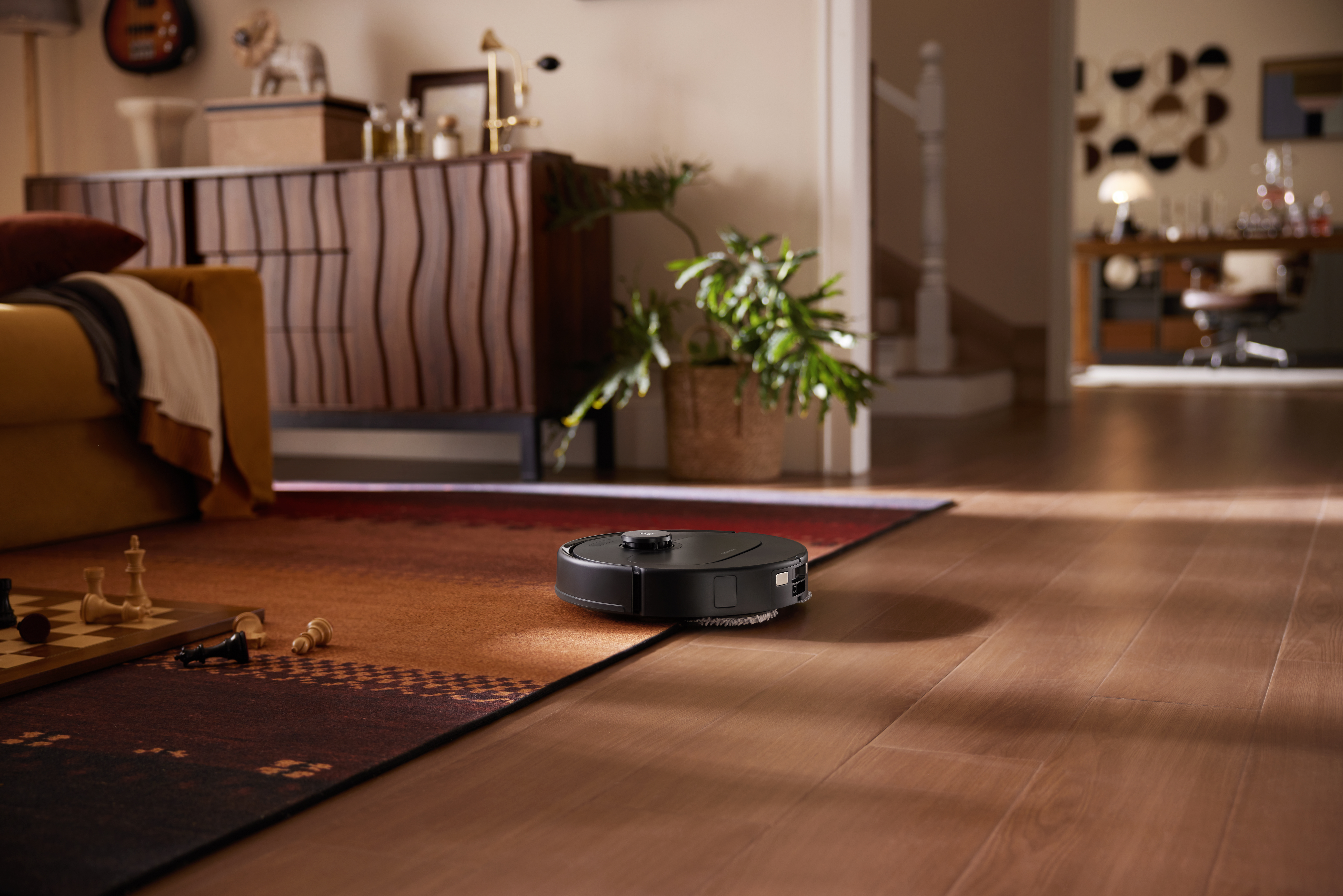 Un robot aspirapolvere nero pulisce un pavimento in legno accanto a un tappeto con pezzi degli scacchi.