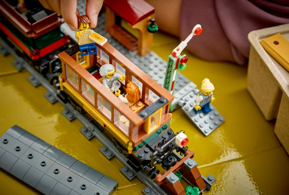 Lego-Zug mit Figuren auf gelbem Untergrund, eine Hand hält ein Ticket.