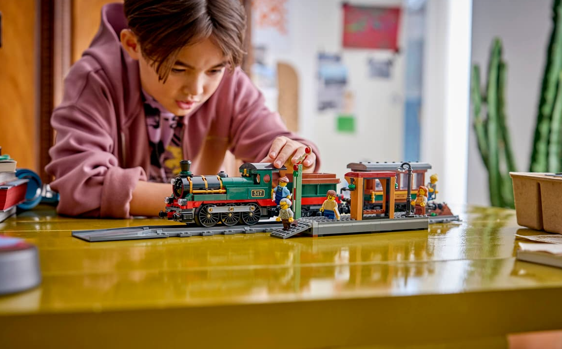Ein Junge baut eine Lego-Eisenbahn mit Bahnhof und Figuren auf einem Tisch.