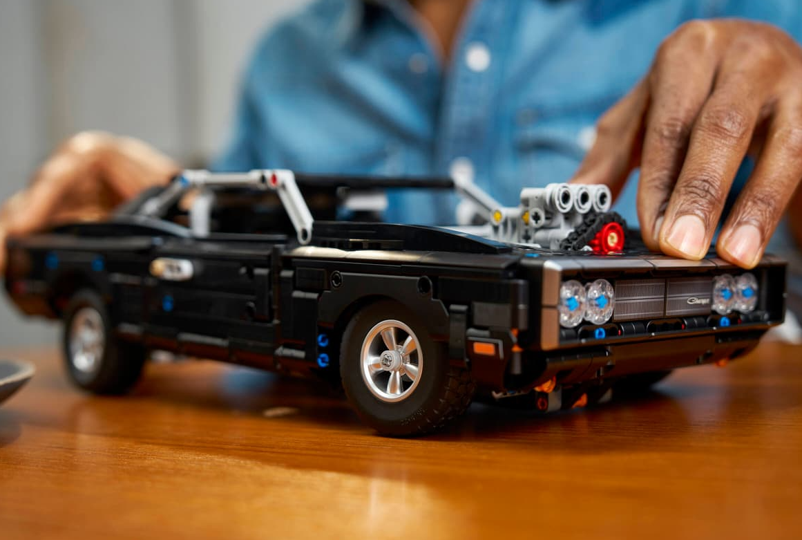 Ein schwarzes Lego-Auto steht auf einem Holztisch, Hände im Hintergrund.