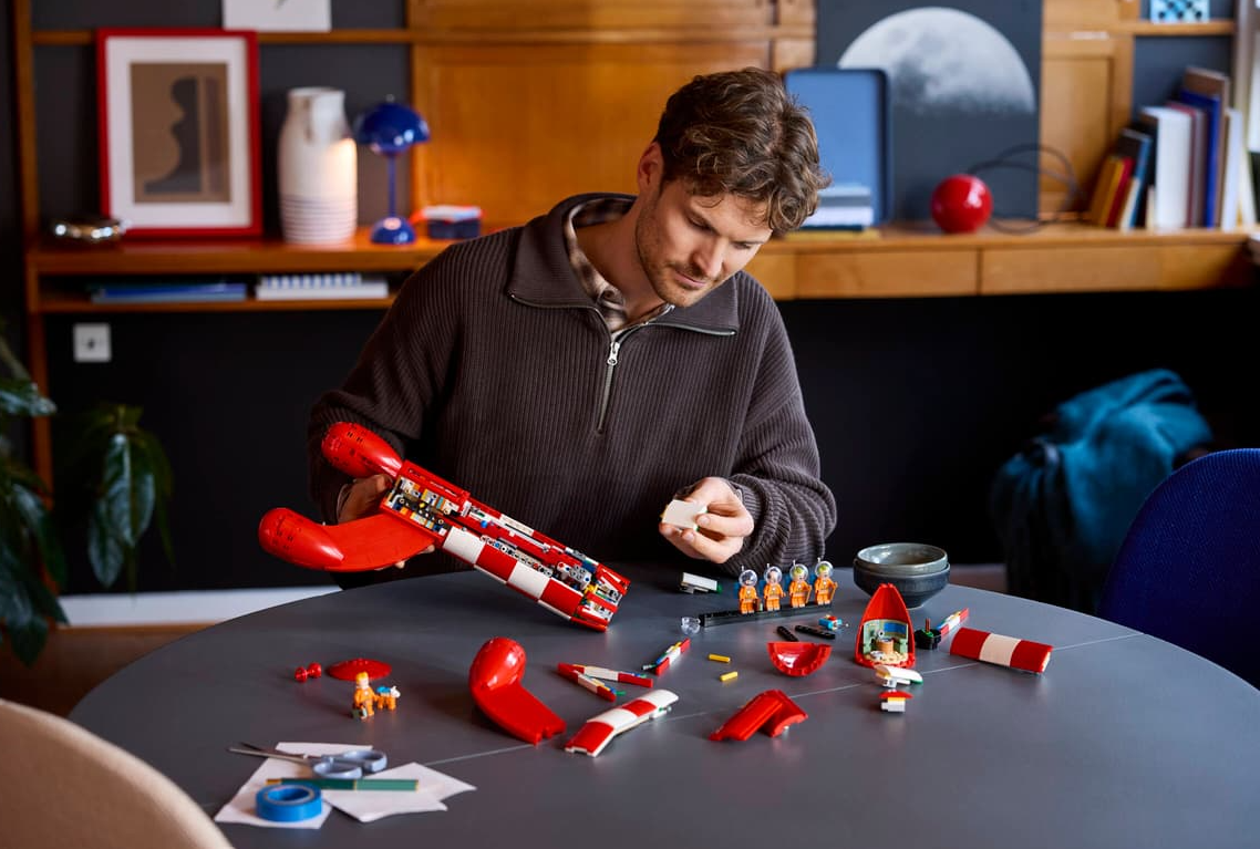 Mann baut roten Lego-Rakete auf einem Tisch mit Astronautenfiguren.