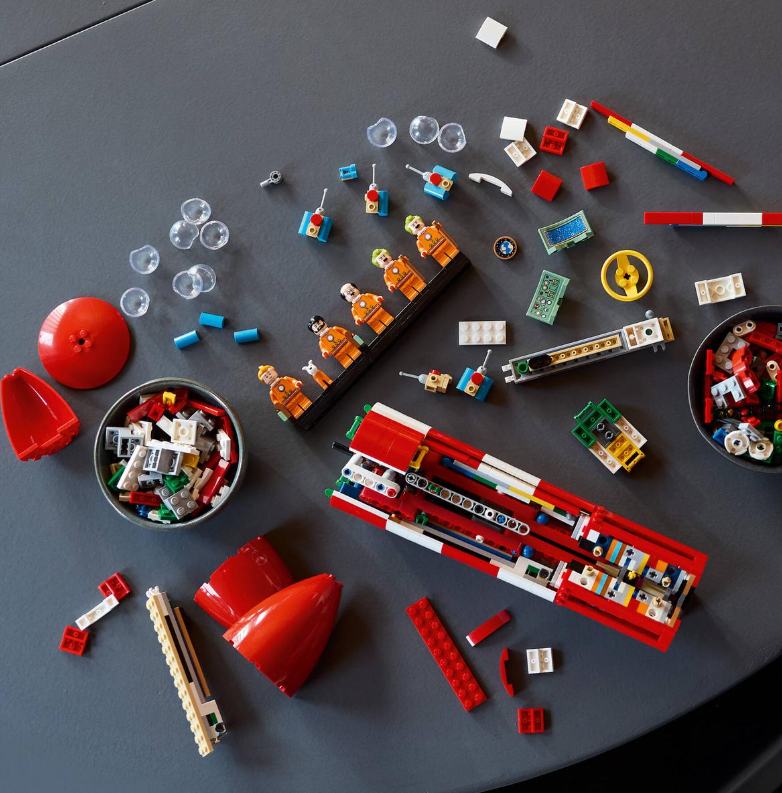 Bunte Lego-Teile liegen auf einem grauen Tisch, darunter Figuren, ein Raketenbausatz und Werkzeuge.