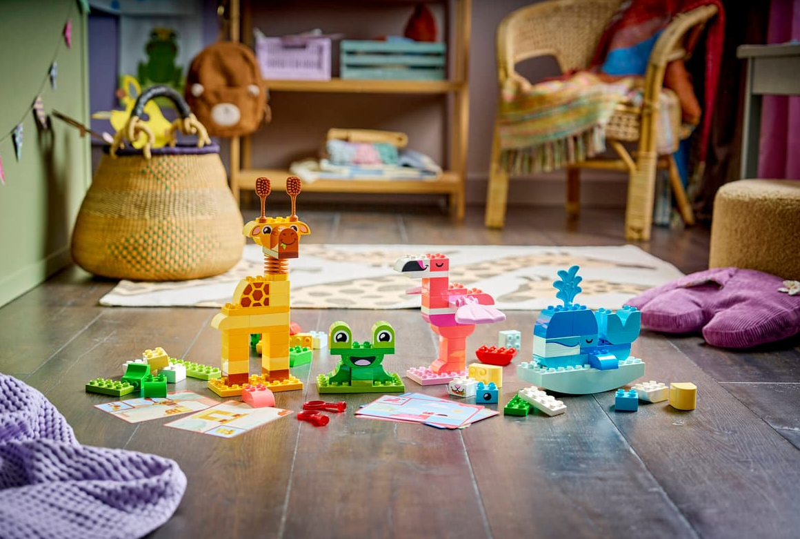 Bunte Lego-Tiere: Giraffe, Frosch, Flamingo, Wal, mit verstreuten Blöcken und Karten auf Holzboden.