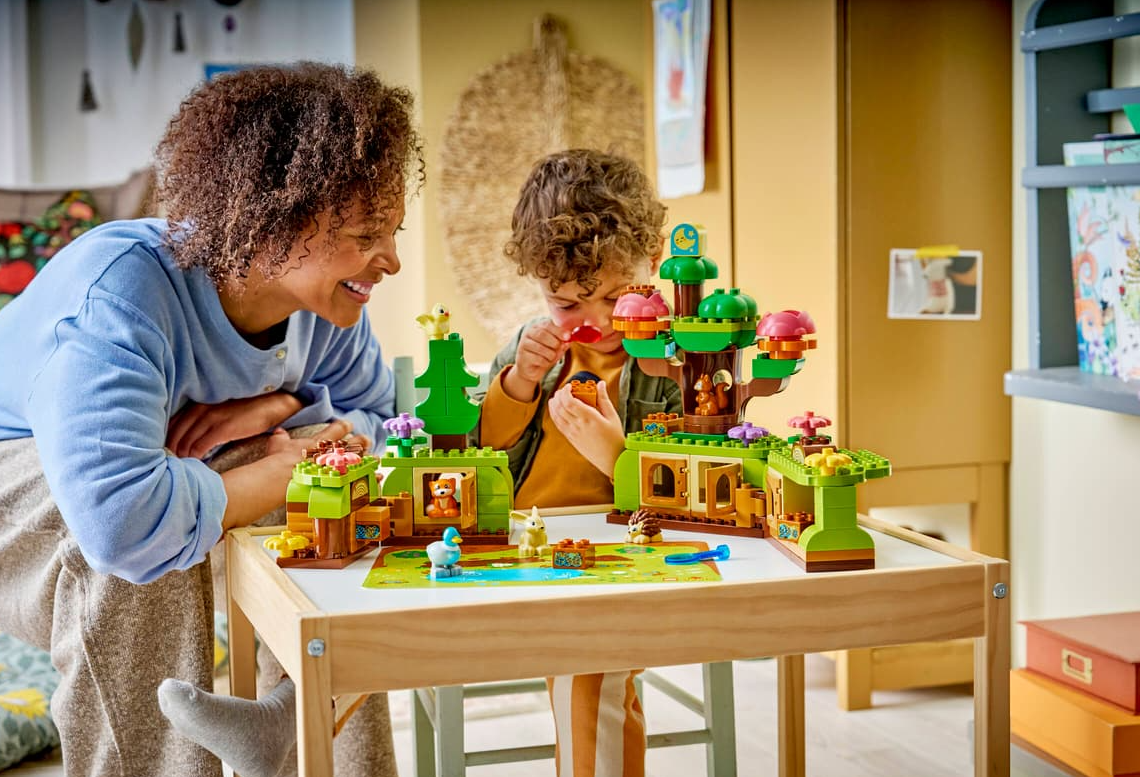 Frau und Kind bauen einen grünen Lego-Wald mit Tieren auf einem Tisch.