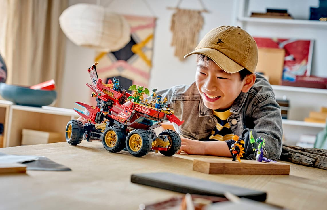 Ein Junge mit Kappe baut ein rotes Lego-Fahrzeug mit Minifiguren und Drachen.