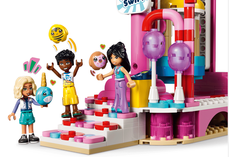 Drei Lego-Figuren spielen mit einem Wal, einem Donut und einer Biene vor einem bunten Gebäude.