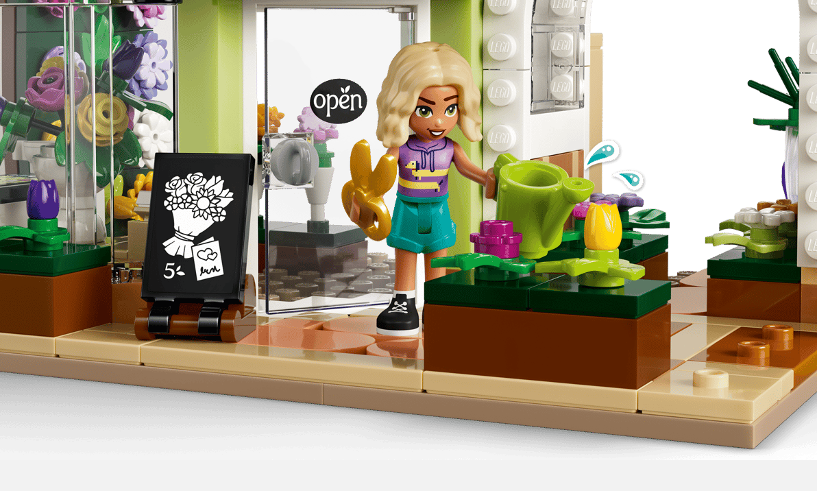 Eine Lego-Figur gießt Blumen in einem Geschäft mit einer "Open"-Tür und einem Blumenarrangement.