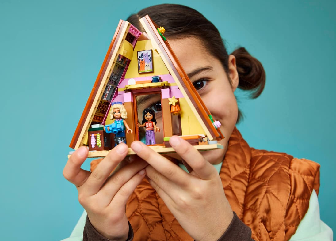 Mädchen hält Lego-Hütte mit zwei Figuren. Blaugrüner Hintergrund.