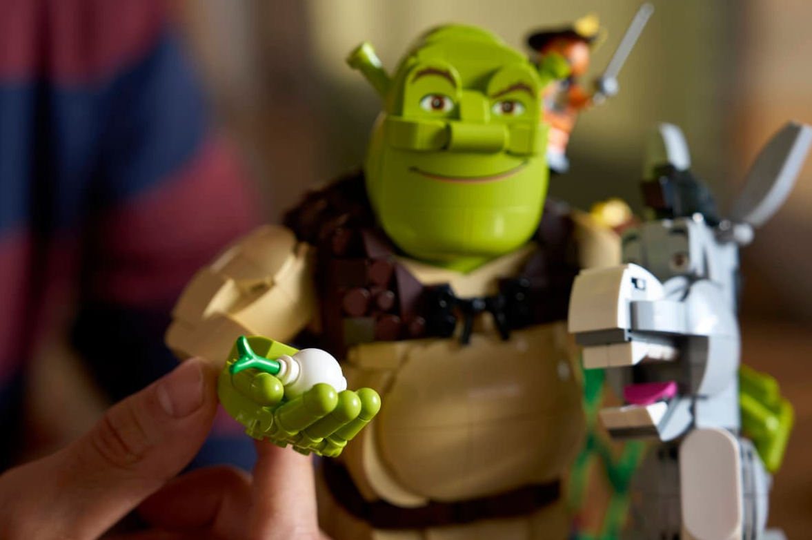 Eine Hand hält ein grünes Lego-Teil mit einem weißen Ei. Shrek und Esel Lego-Figuren im Hintergrund.