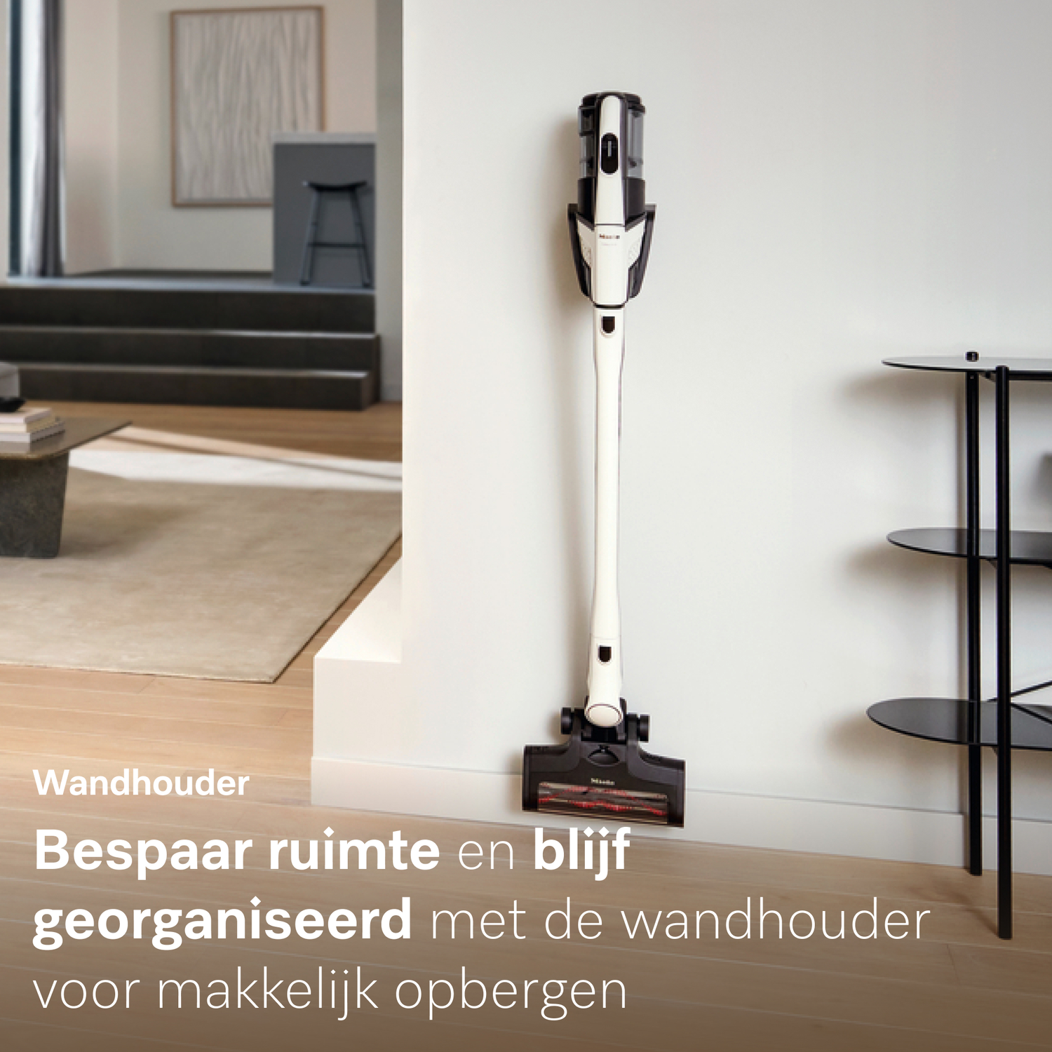 Een witte steelstofzuiger hangt aan een muurbeugel. Rechts staat een rekje.