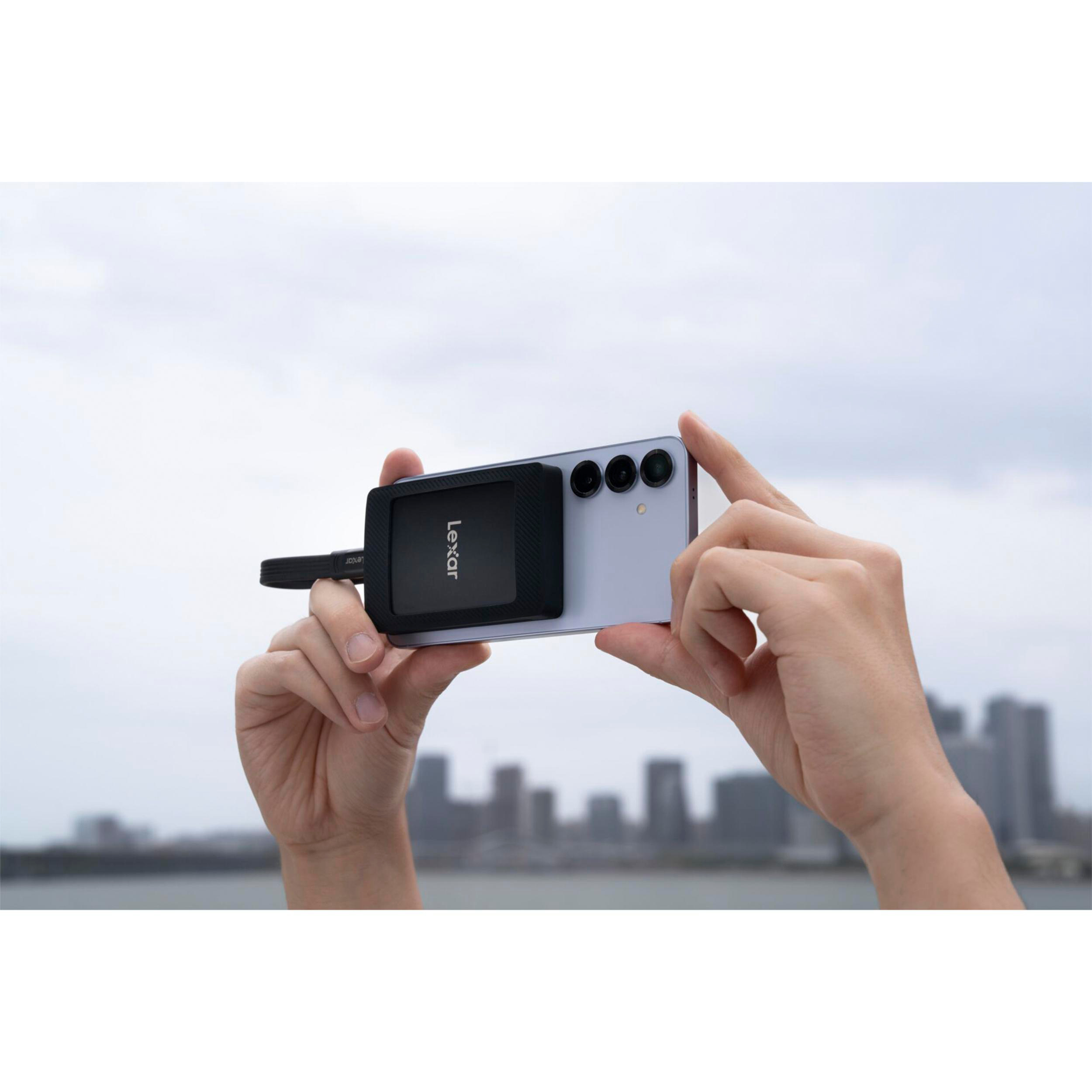 Eine Person hält ein Smartphone mit angebrachtem Lexar-SSD-Laufwerk. Wolkenverhangener Himmel.