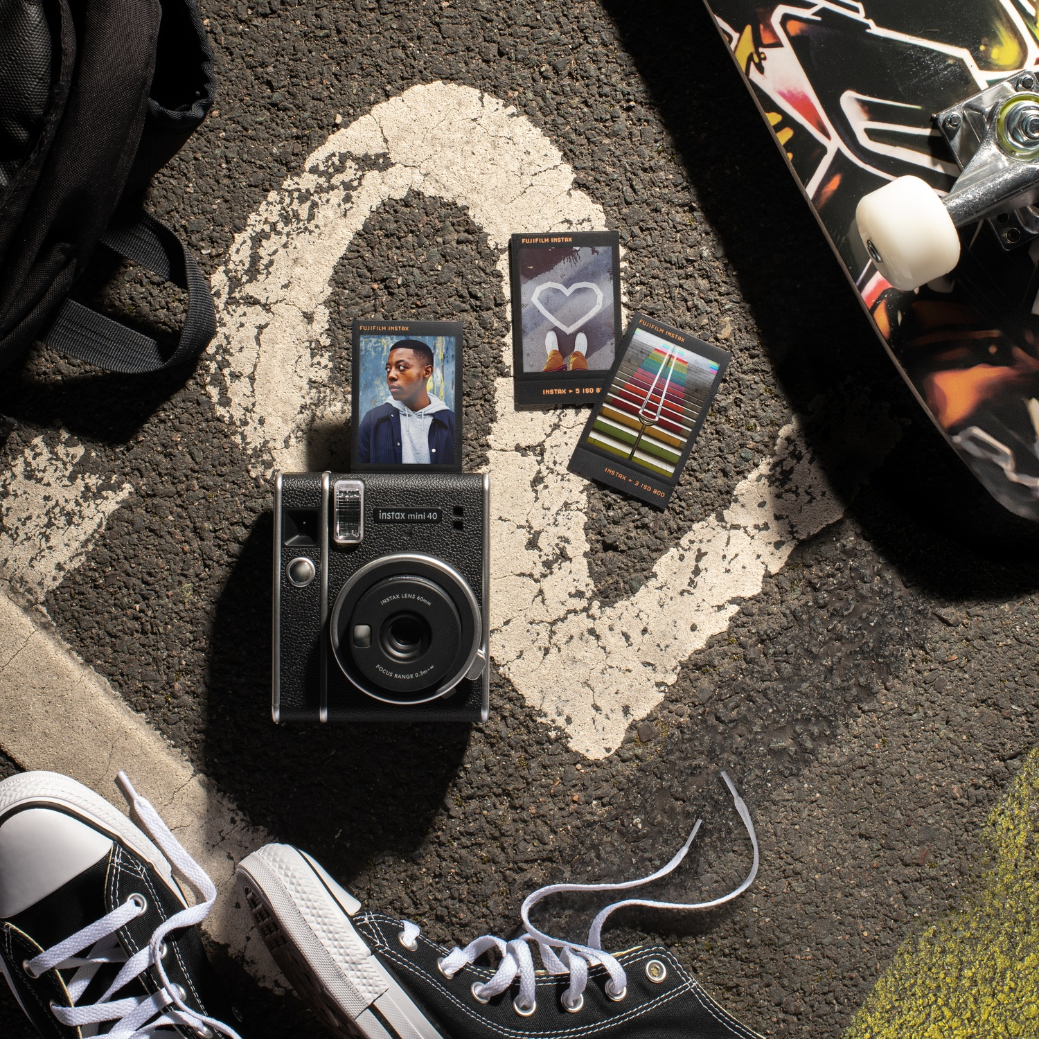Een Instax Mini 40 camera, skateboard, rugzak en sneakers op asfalt.