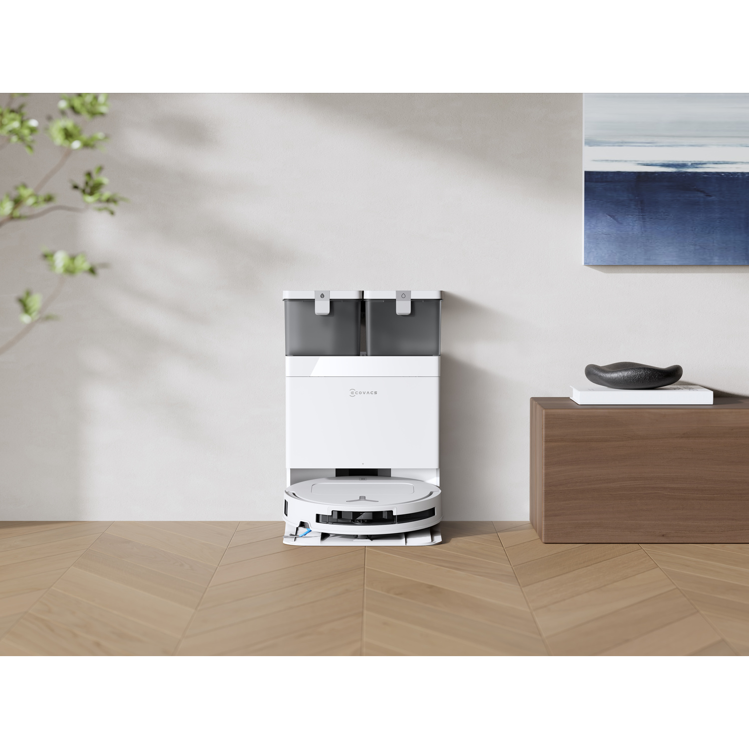 Un robot aspirapolvere bianco con stazione di svuotamento automatico si trova su un pavimento in legno, contro una parete bianca con un quadro blu.