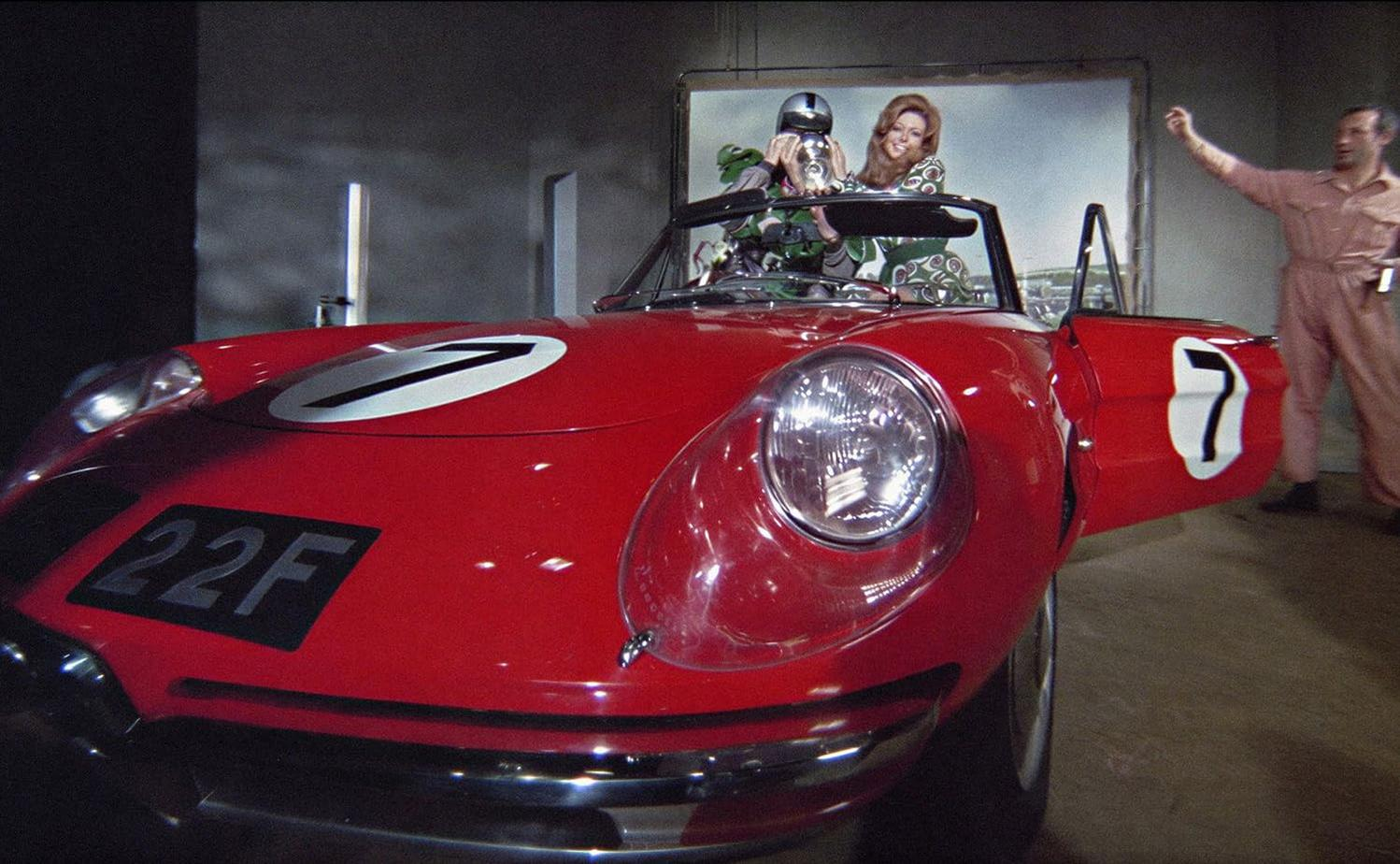 Ein rotes Rennauto mit zwei Frauen und einem Mann im Hintergrund.