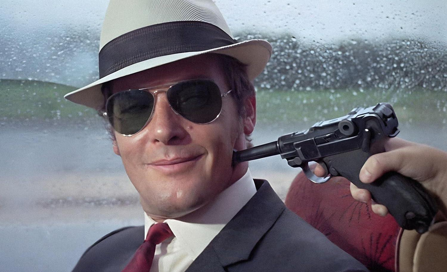 Ein Mann im Anzug mit Hut und Sonnenbrille, dem eine Pistole an die Wange gehalten wird.