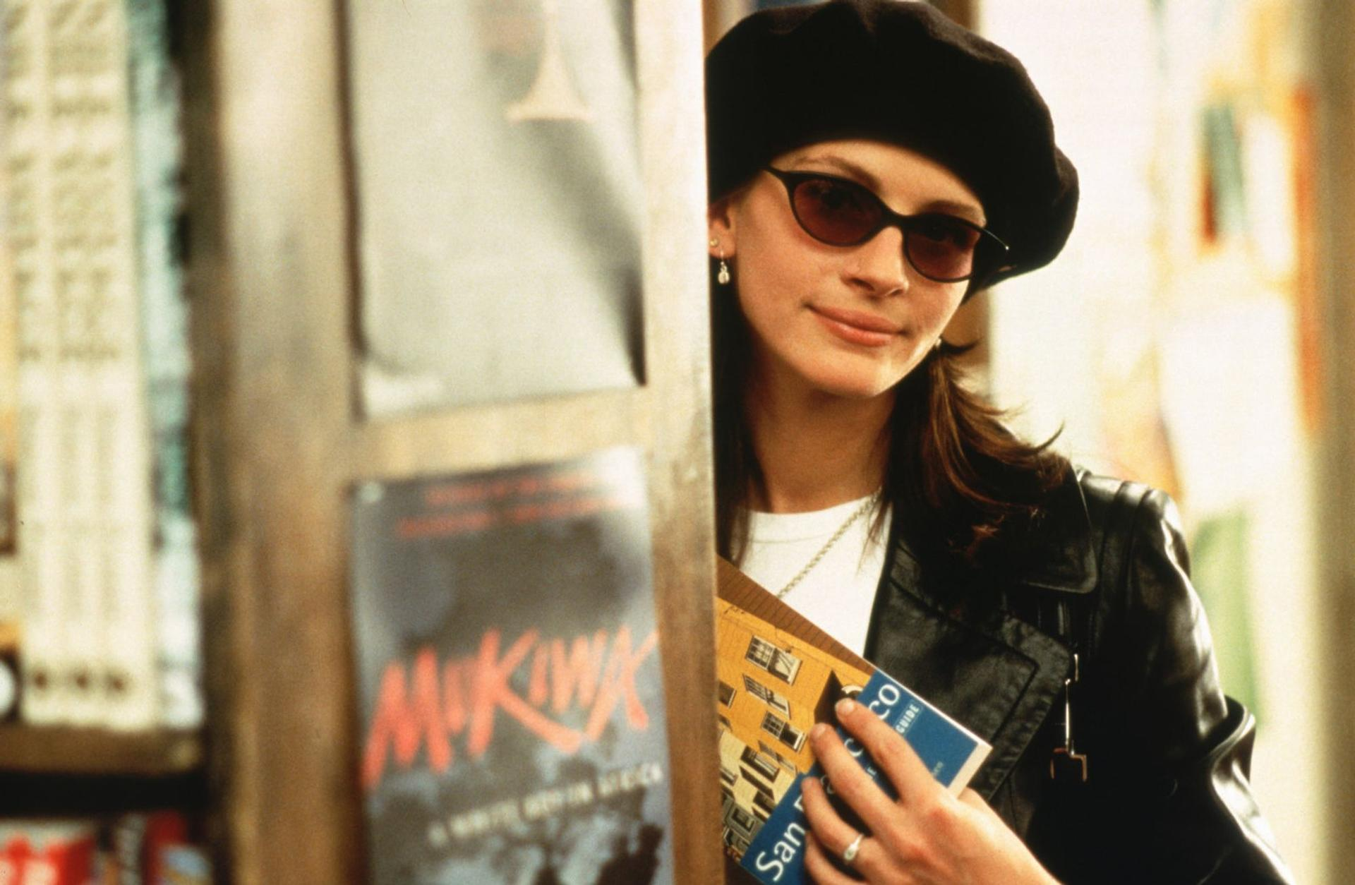 Eine Frau mit schwarzem Barett und Sonnenbrille hält Bücher vor einem Regal.