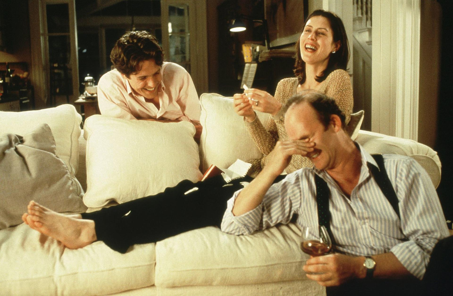 Drei Personen lachen auf einem Sofa. Ein Mann liegt, eine Frau sitzt, ein Mann sitzt und hält ein Glas Wein.