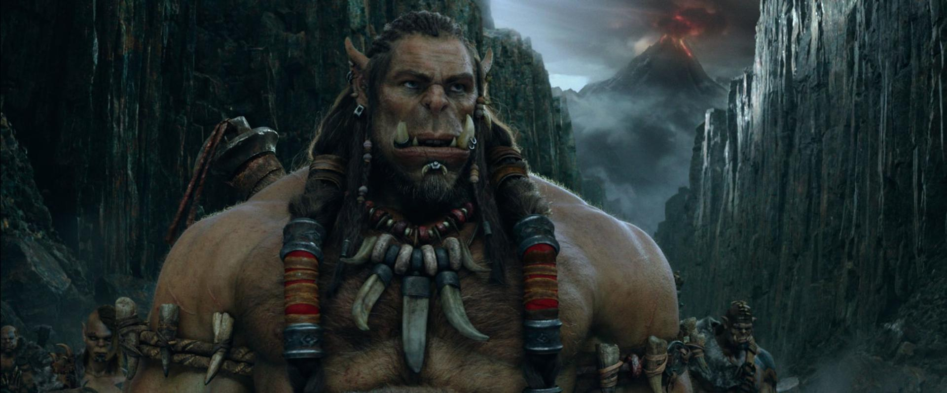 Ein großer Ork mit langen Haaren und einem Knochenhalsband steht vor einer Berglandschaft.