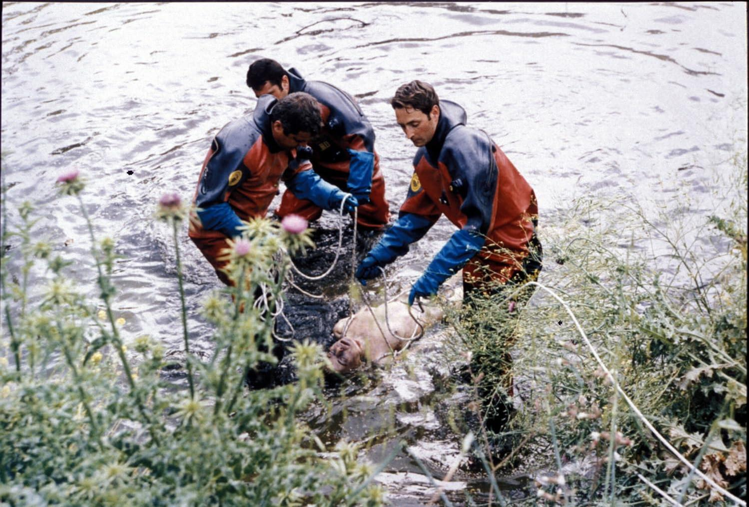 Drei Taucher in roten Anzügen bergen eine Person aus einem Fluss.