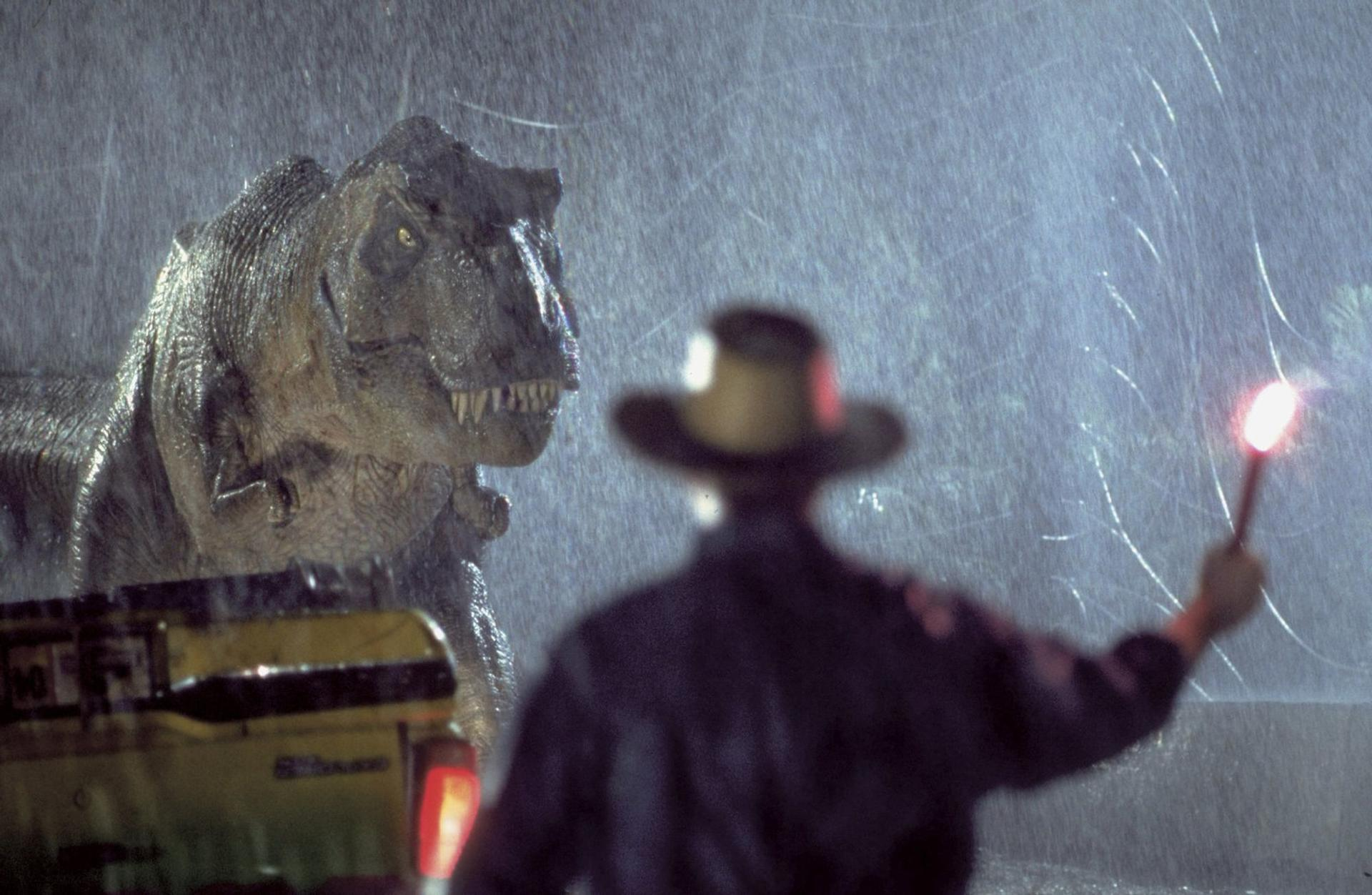 Ein T-Rex brüllt im Regen neben einem gelben Fahrzeug, während eine Person mit Fackel im Vordergrund steht.