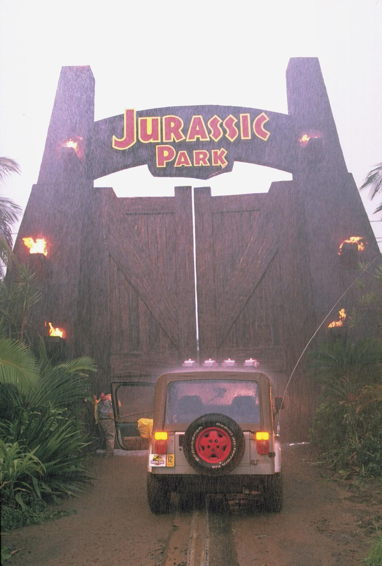 Ein Jeep fährt auf ein Tor mit der Aufschrift Jurassic Park zu. Es regnet.