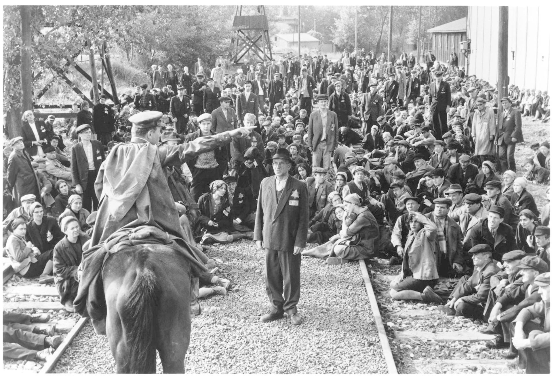 Eine Gruppe von Menschen sitzt und steht auf Bahngleisen, ein Mann zu Pferd blickt auf.