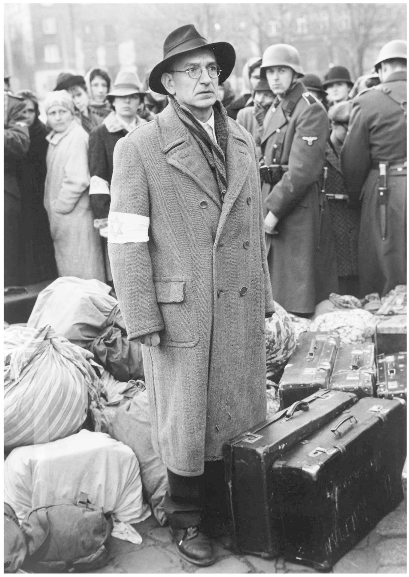 Ein Mann mit Hut und Brille trägt ein Band mit Stern. Im Hintergrund stehen Menschen und Soldaten.