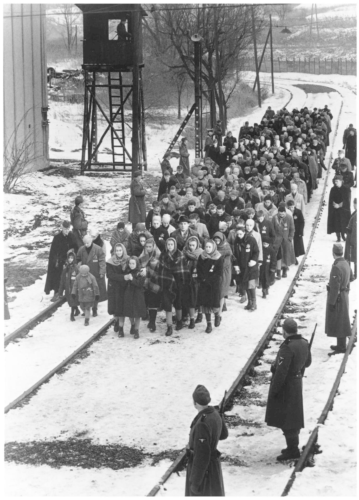 Gefangene marschieren im Schnee an Bahngleisen, bewacht von Soldaten mit einem Wachturm im Hintergrund.