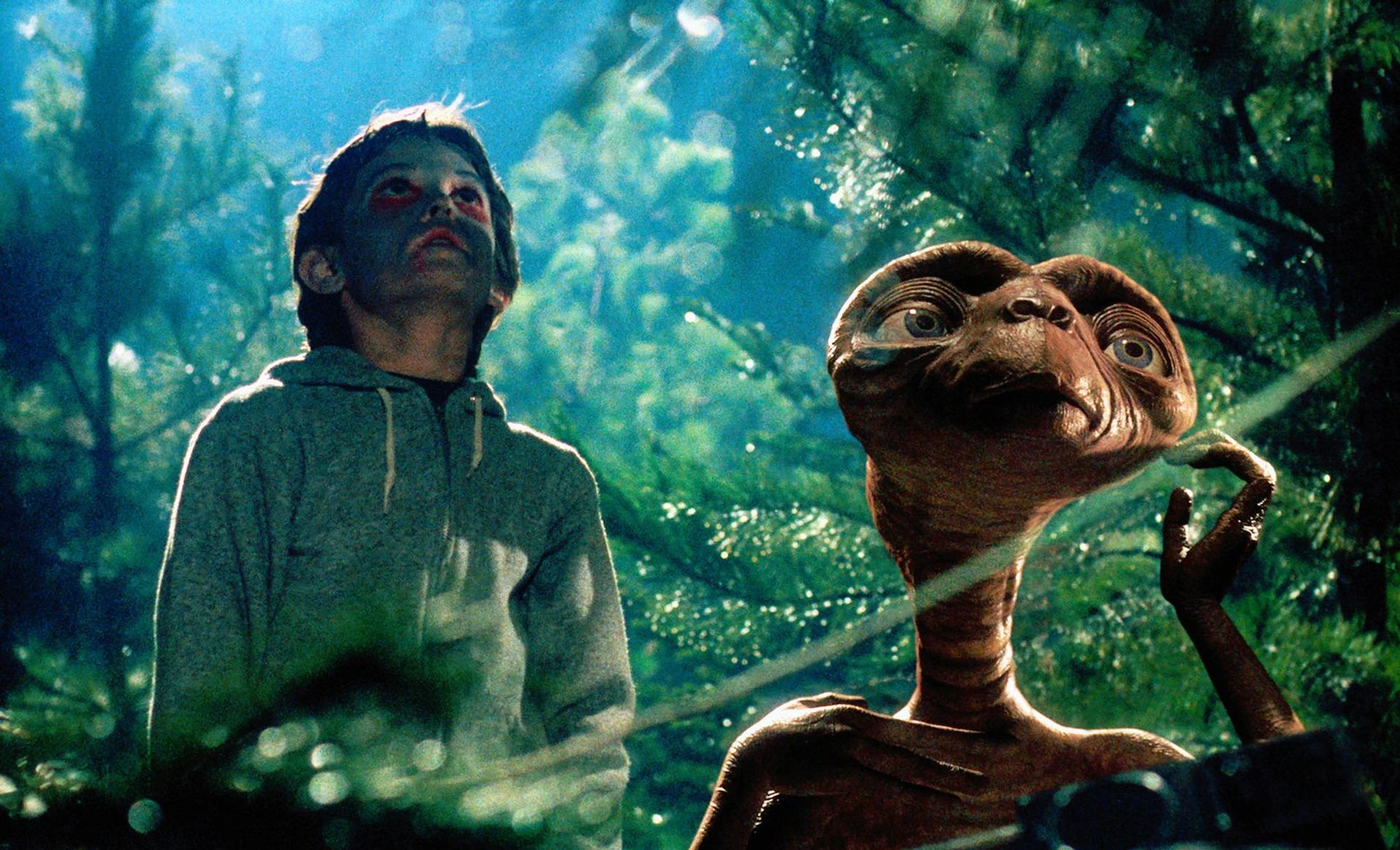 Ein Junge mit dunkler Schminke im Gesicht und E.T. stehen im Wald.