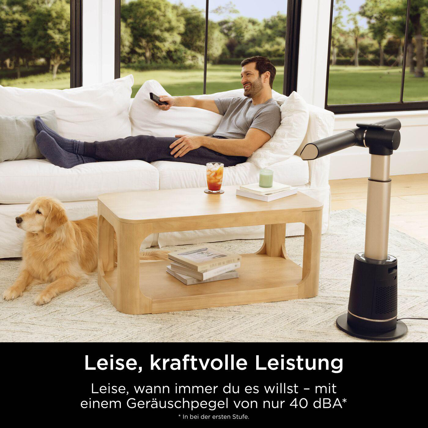 Mann auf Sofa mit Hund, Tisch, Ventilator und Getränk. Draußen Bäume.