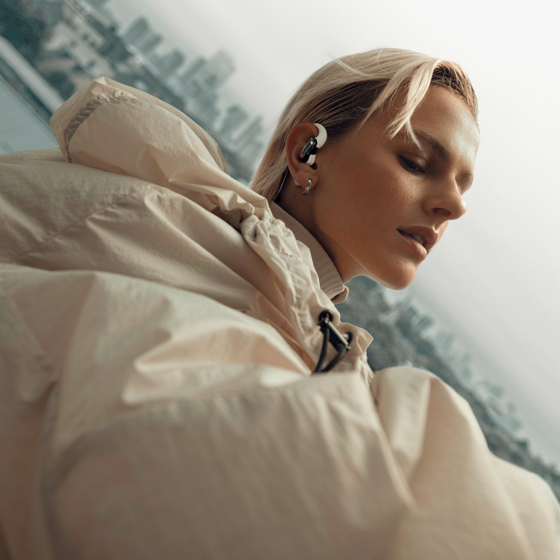 Blonde Frau mit Ohrhörern trägt eine beige Jacke, mit Stadt im Hintergrund.