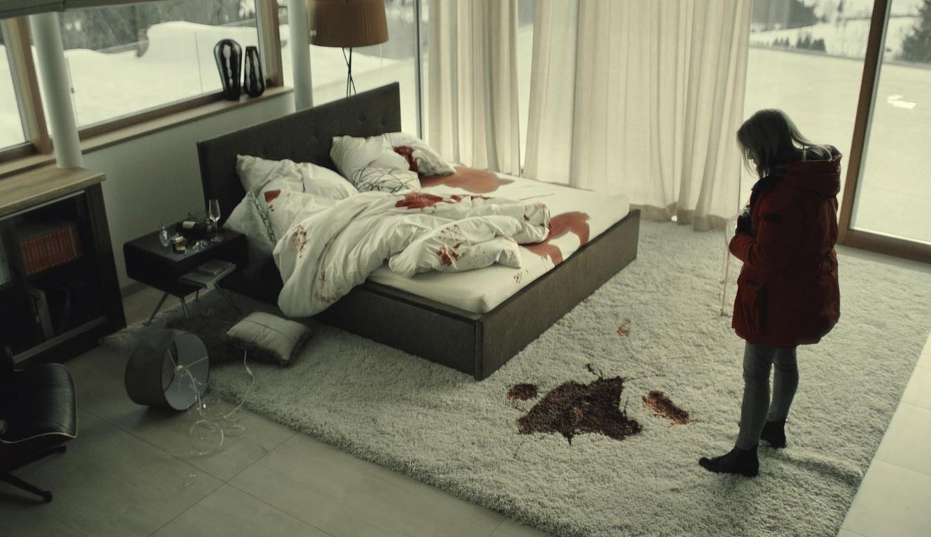 Ein Mädchen in rotem Mantel steht neben einem Bett mit Blutspuren.