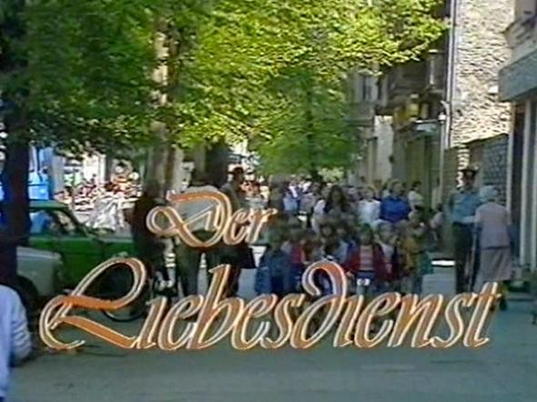 Eine Straßenszene mit dem Titel „Der Liebesdienst“ und vielen Menschen auf einem Gehweg.