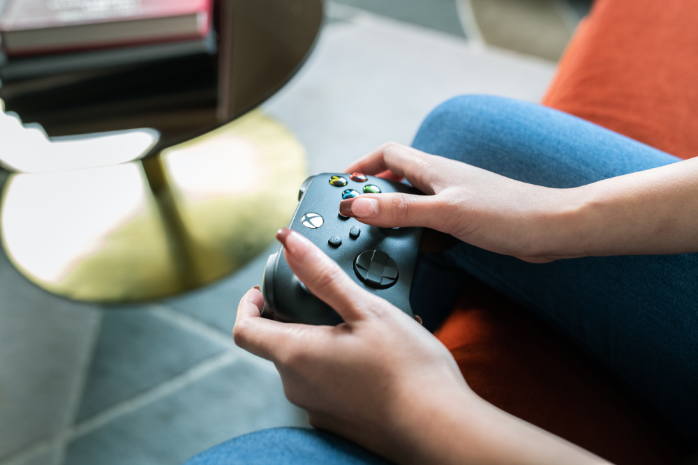 Manos sujetando un mando negro de Xbox, con vaqueros azules. Sentado en una silla naranja.