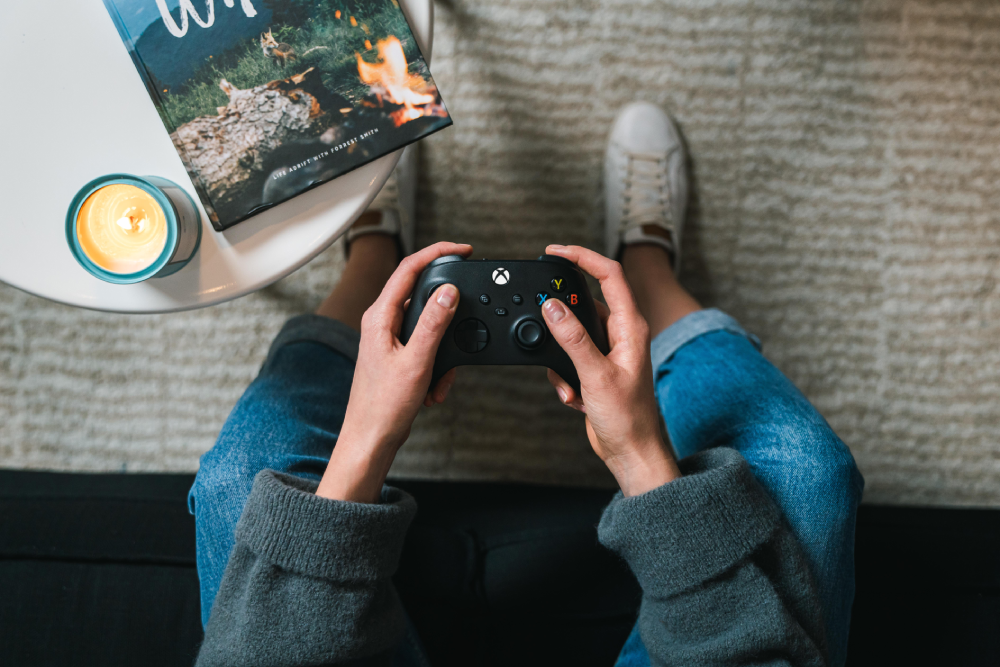 Una persona sostiene un mando negro de Xbox, sentada en una alfombra con vaqueros.
