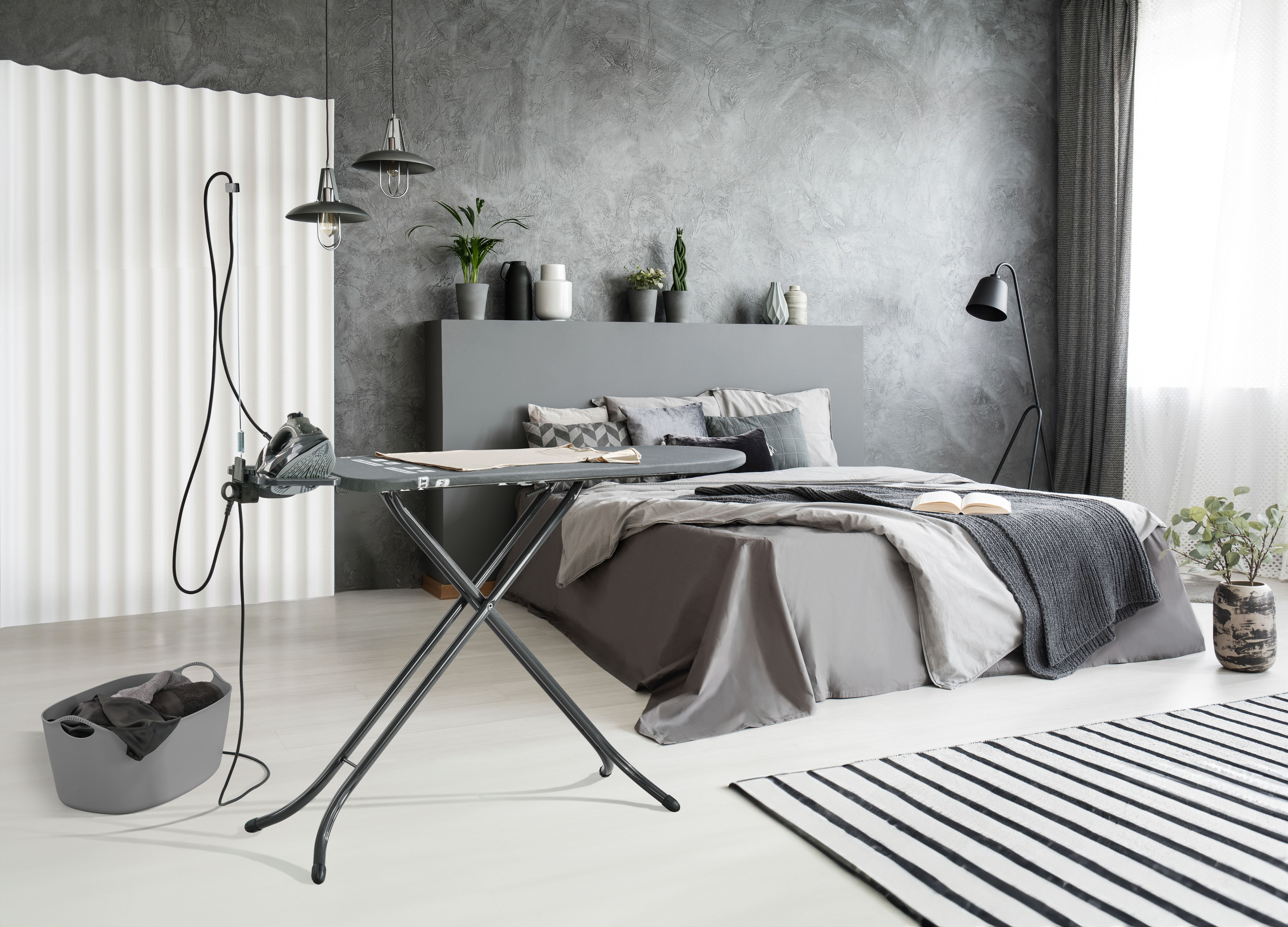 Ein Schlafzimmer mit einem Bügelbrett und Bügeleisen im Vordergrund, daneben ein Korb.