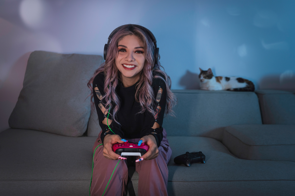 Una mujer con pelo morado juega videojuegos en un sofá, un gato descansa al fondo.