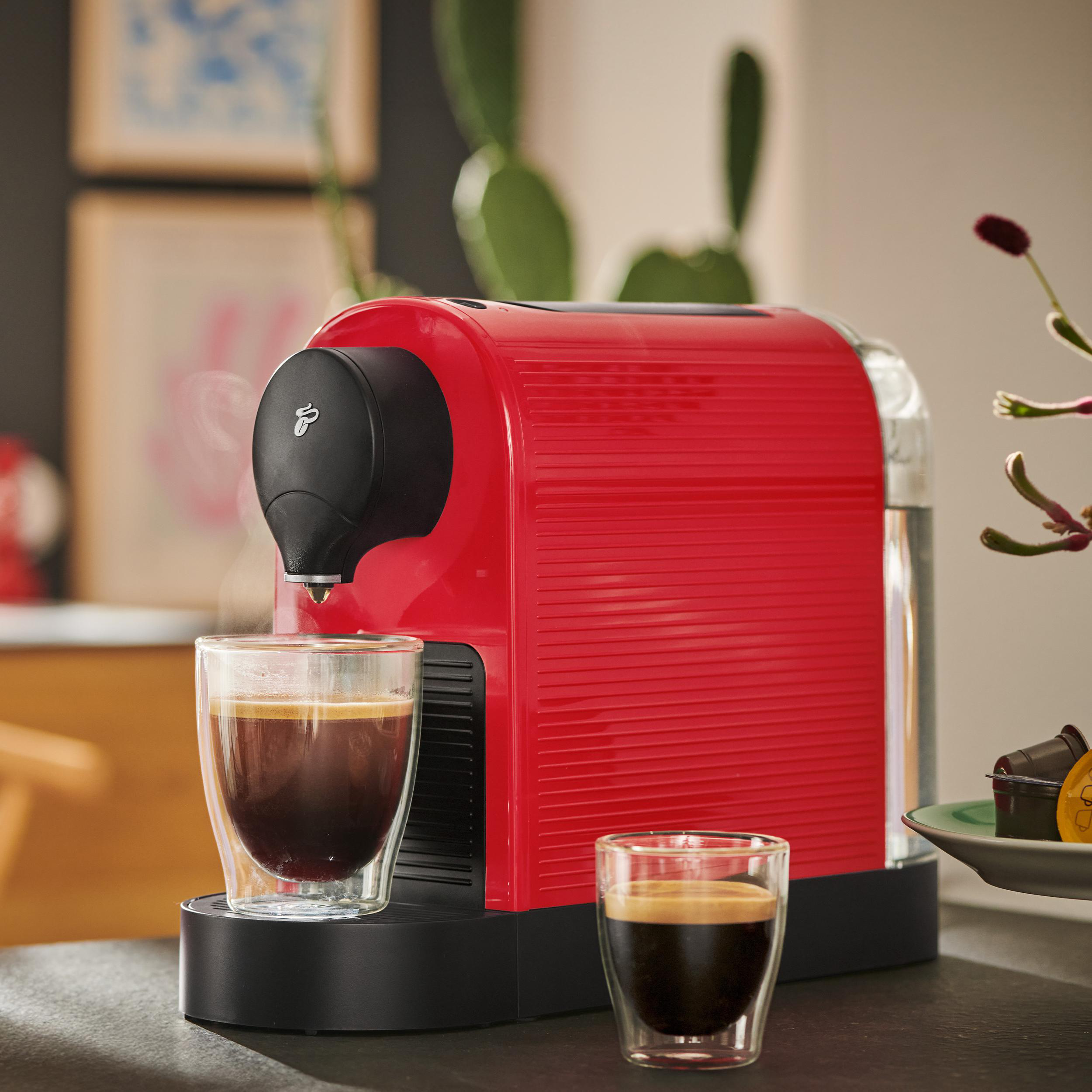 Eine rote Kaffeemaschine brüht Kaffee in zwei Gläser. Im Hintergrund sind Pflanzen.