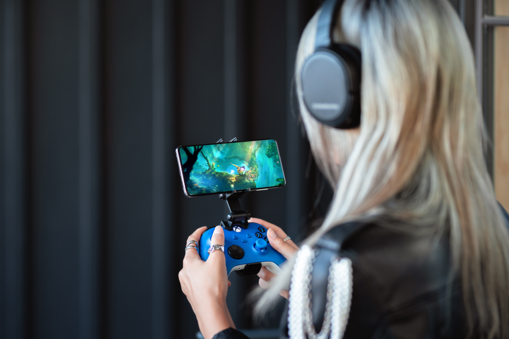 Una mujer con auriculares juega un videojuego en su teléfono con un mando azul.