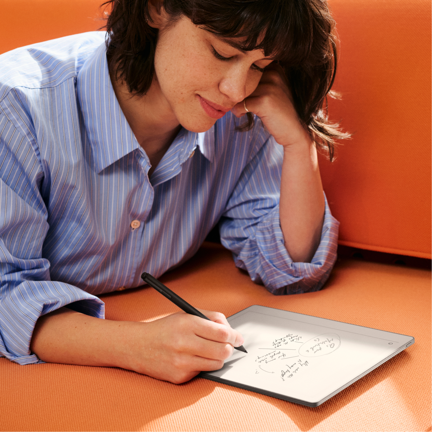 A woman in a blue striped shirt writes on a tablet while resting on an orange couch.