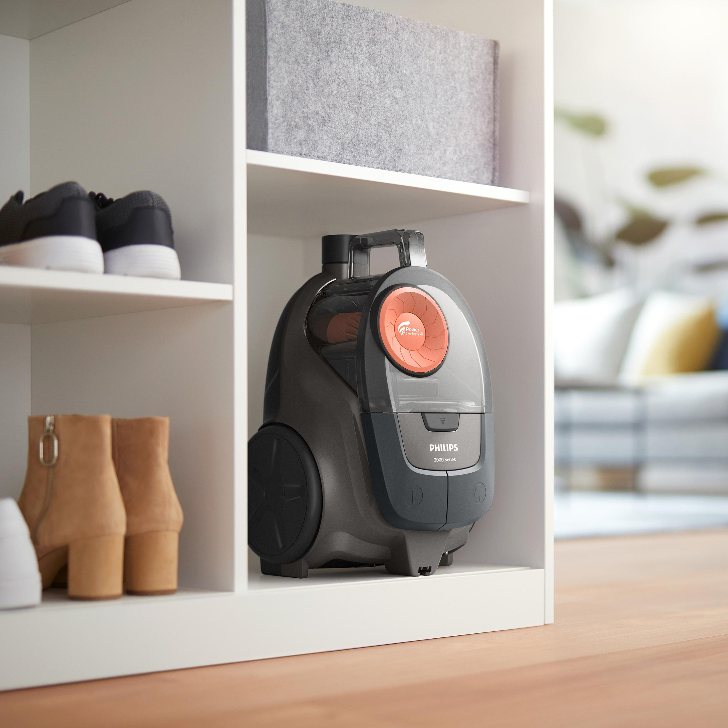 Grauer Philips Staubsauger mit orangefarbenem Detail auf einem Regal, neben Schuhen.