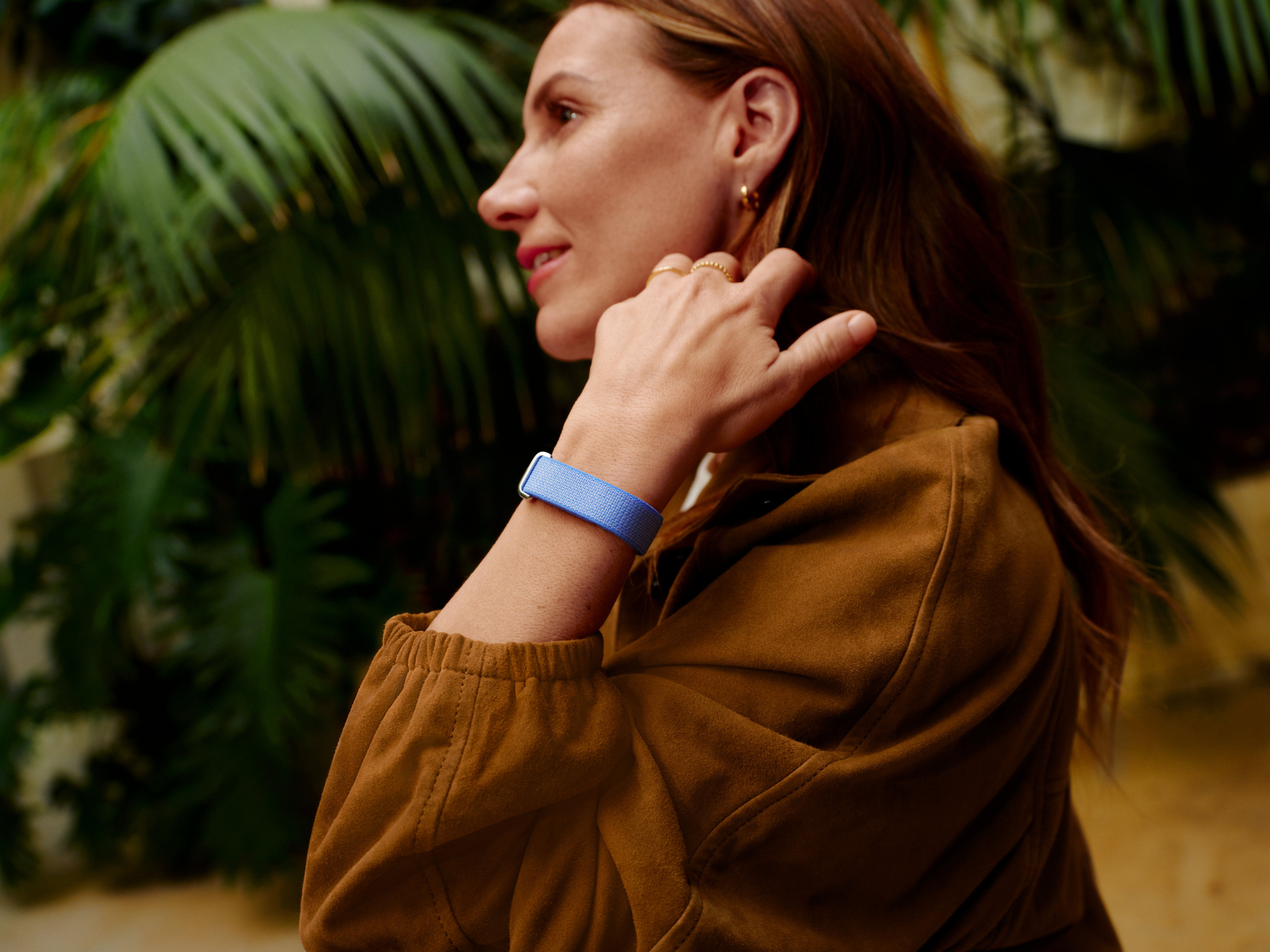 Frau mit brauner Jacke und blauem Armband, im Hintergrund grüne Blätter.
