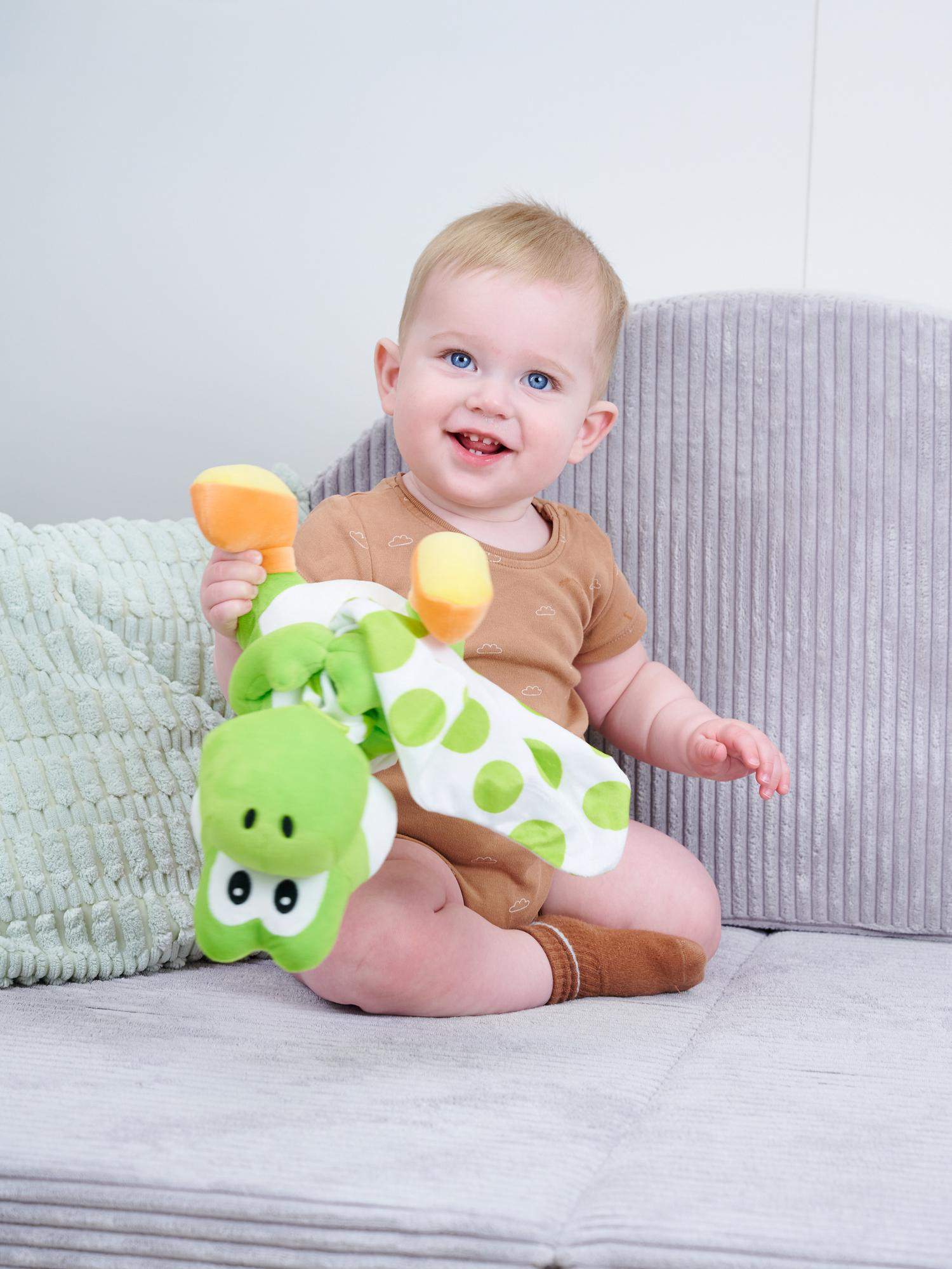Ein Baby mit blauen Augen sitzt auf einem Sofa und hält eine grüne Krokodil-Spielzeug.