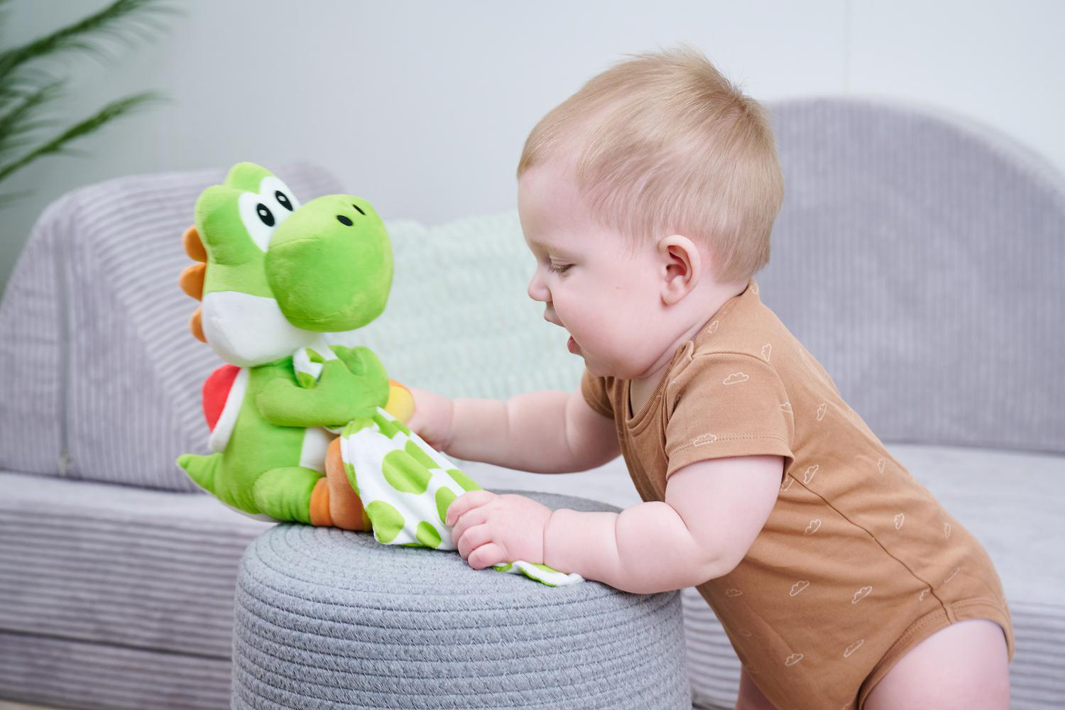 Ein Baby in brauner Kleidung spielt mit einer grünen Yoshi-Puppe auf einem Hocker.