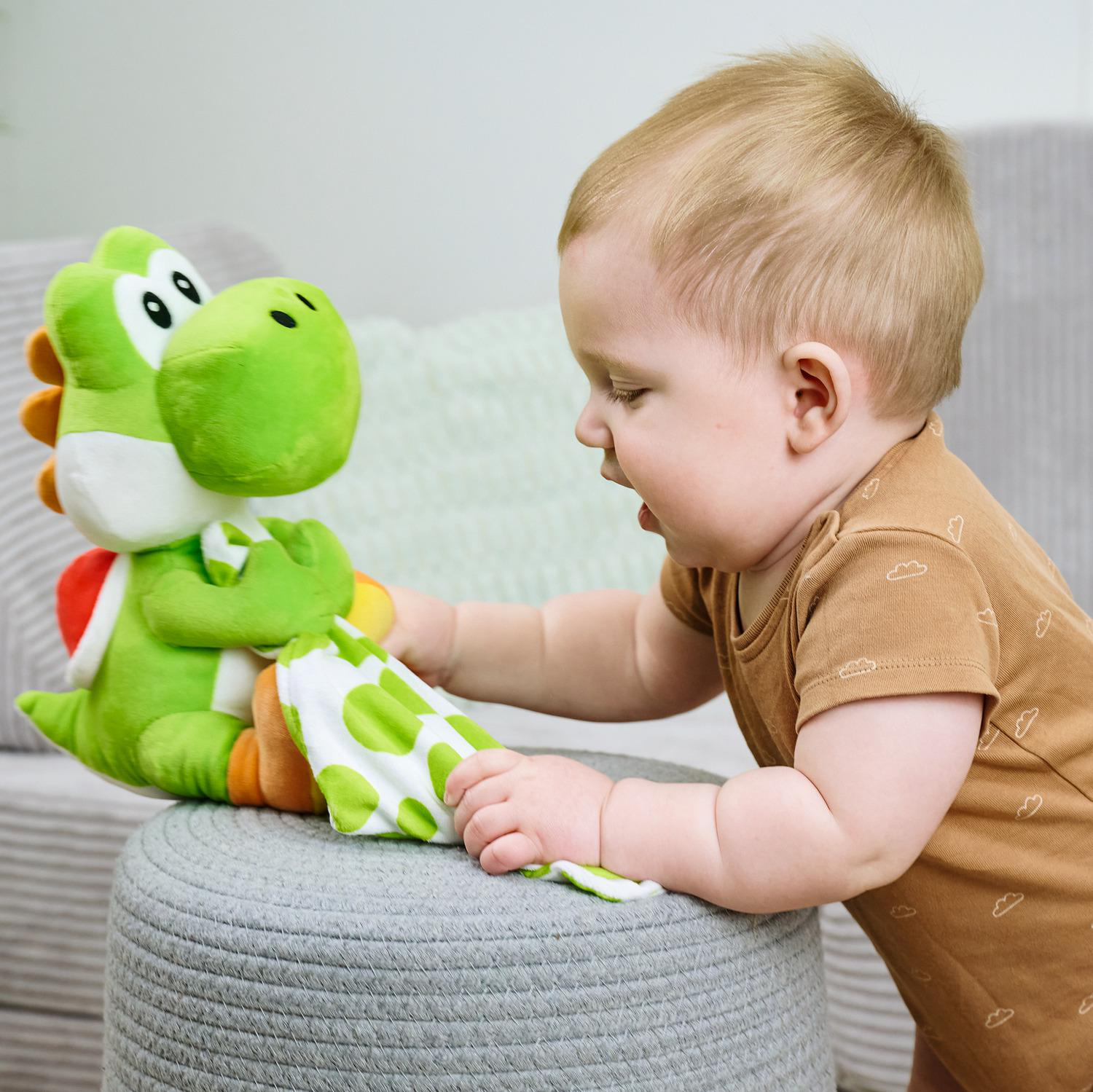 Ein Baby spielt mit einem grünen Yoshi-Plüschtier auf einem grauen Hocker.