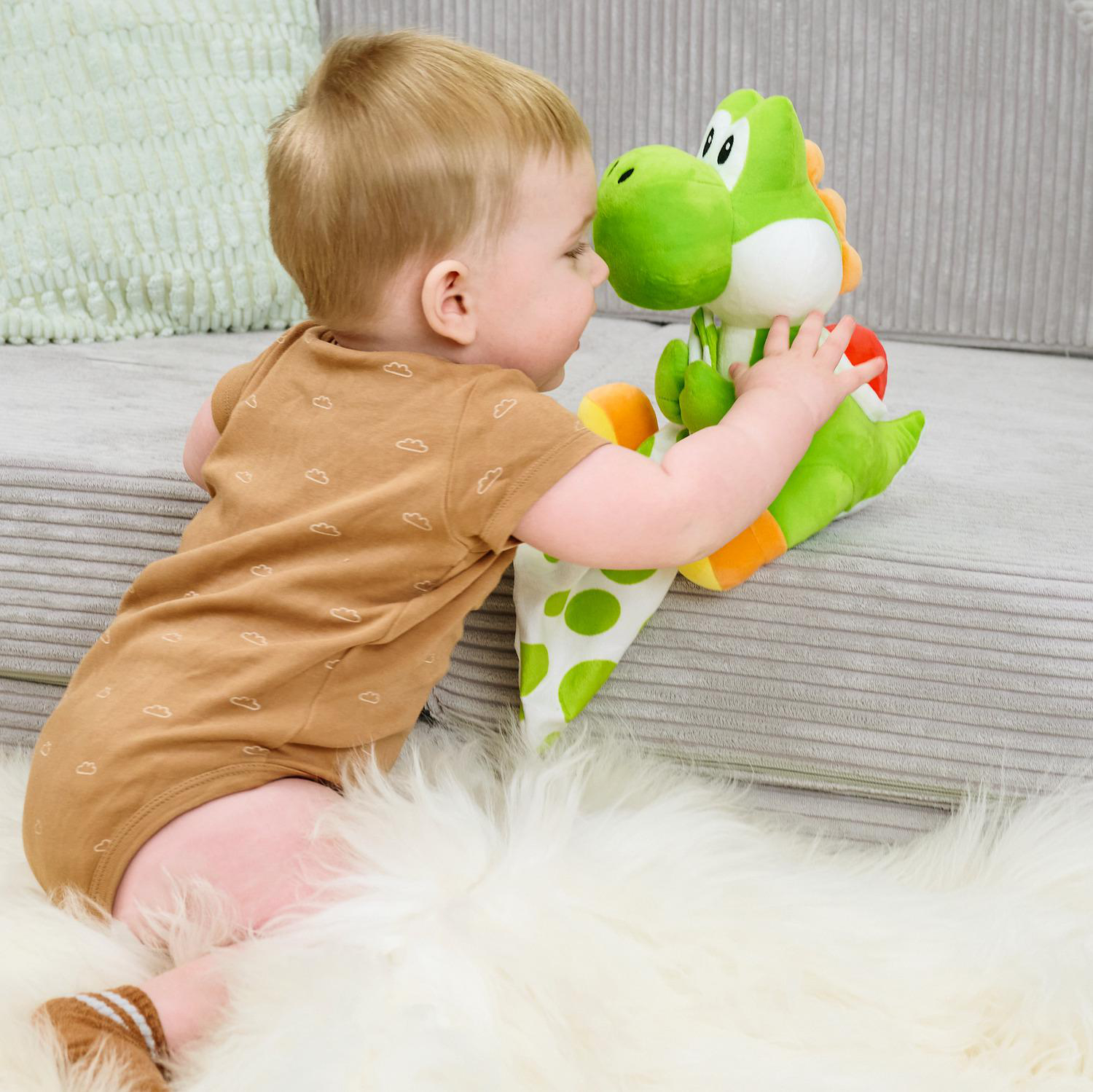 Ein Baby in einem braunen Strampler spielt mit einer grünen Yoshi-Puppe auf einer weißen Decke.