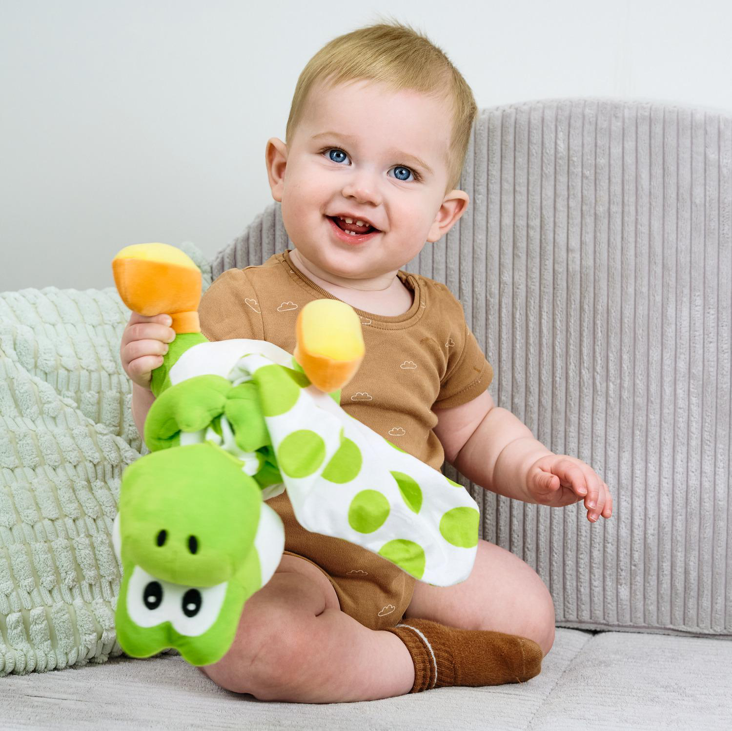 Ein Baby sitzt und hält ein grünes Stofftier mit Tupfen. Es trägt braune Kleidung.