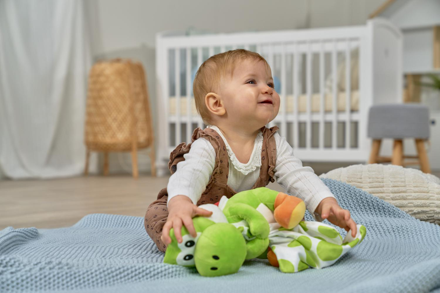 Ein Baby in einem weißen Hemd und braunen Latzhosen spielt mit einem grünen Plüschtier auf einer blauen Decke.